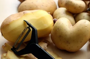 Zemiaky - jeden čiastočne očistený zemiak a škrabka na pozadí zemiakov