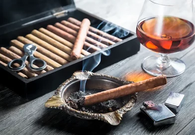Cigary - príslušenstvo: humidor naplnený cigarami, kovový orezávač, horiaca cigara v kovovom popolníku, zapaľovač a pohár s likérom