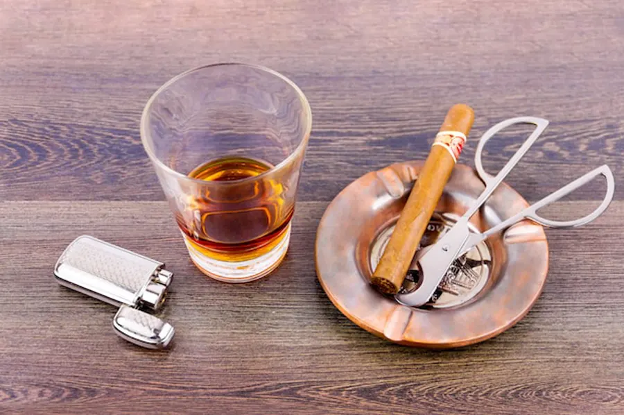 Cigarové príslušenstvo: zapaľovač, pohár s whisky, cigara a orezávač v popolníku