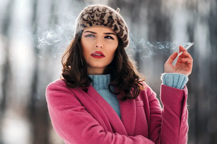 Mladá žena v ružovom kabáte a vzorovanej baretke fajčí cigaretu
