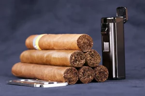 Päť cigár poskladaných na kôpku, tryskový zapaľovač a kovový orezávač na cigary