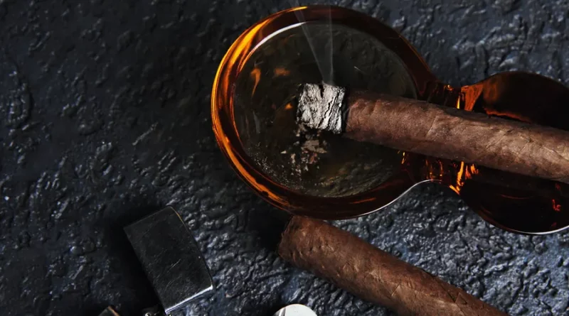 Horiaca cigara odložená v popolníku, cigarový zapaľovač a nezapálené cigara na tmavom pozadí