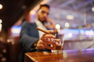 Detail mužskej ruky, ktorá drží cigaru a pohár s alkoholom, na pozadí mladý muž v obleku