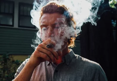Mladý muž s dlhšími vlasami fajčí ciguru, tvár mu zahaľuje hustý dym