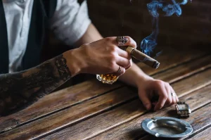 Mladý muž fajčí cigaru a drží v ruke pohárik s alkoholom - detail potetovaných rúk mladého muža